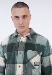 Man met kort haar in een groen-wit geruit overhemd met knopen, voorzien van een borstzak en een "Protest" logo patch.