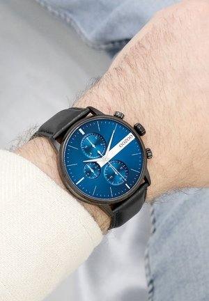 Montre chronographe avec un cadran bleu, un boîtier noir et un bracelet en cuir. Présente trois sous-cadrans, des accents argentés et des index horaires blancs.