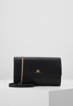 Czarna skórzana kopertówka Emporio Armani z złotym łańcuszkiem i logo, spoczywająca na białej powierzchni ekspozycyjnej na jednolitym szarym tle.