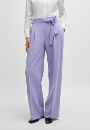 Vrouw met hooggetailleerde, wijde lavendelkleurige broek met een gestrikte riem, gecombineerd met een wit overhemd en zwarte puntige schoenen.