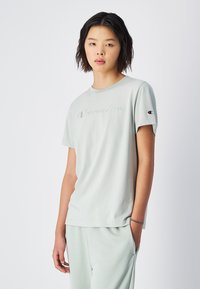 T-shirt en coton vert clair avec un logo "Champion" en relief sur le devant et un petit patch logo sur la manche gauche. Manches courtes, col rond.