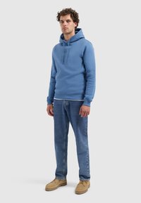 Blauwe capuchontrui met een buidelzak en een koord, gecombineerd met lichtblauwe jeans en tan schoenen. De textuur lijkt zacht en casual.