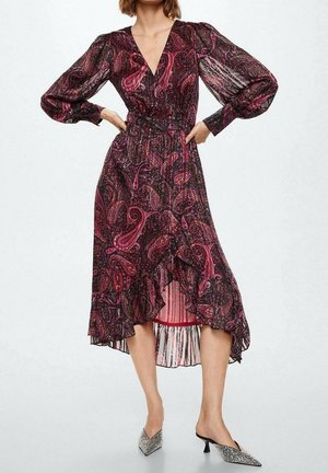 Frau trägt ein langärmliges, V-Ausschnitt, knielanges Kleid mit rosa-schwarzem Paisley-Muster und Rüschensaum, kombiniert mit spitzen Kitten-Heel-Schuhen.