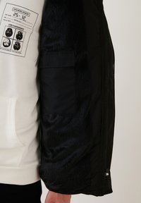 Sort jakke med en tekstureret, pelsagtig inderside og mat yderside, udstyret med en sidelomme og en hvid sweatshirt med trykte detaljer.