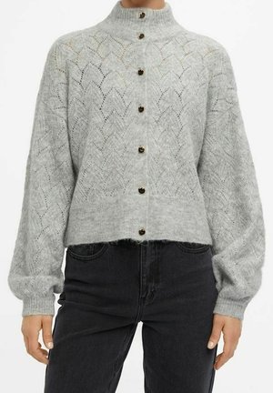 Jasnoszary dzianinowy kardigan z koronkowym wzorem, wysokim kołnierzem, bufkami na długich rękawach oraz sześcioma ciemnymi guzikami z przodu, noszony z czarnymi dżinsami.