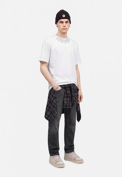 Jeune homme portant un t-shirt blanc avec texte, un bonnet noir avec un smiley jaune, une chemise à carreaux nouée autour de la taille, un jean noir et des baskets beiges.