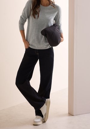 Žena v šedém tričku s dlouhým rukávem, černých širokých kalhotách, bílých teniskách, držící černou péřovou bundu, stojící uvnitř.