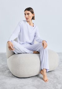 Svetlo vijoličen komplet za prosti čas z dolgo majico z gumbki na izrezu in hlačami sproščenega kroja, oblečen med sedenjem na kremni stolk z teksturo.