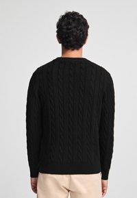 Svart stickad tröja med kabelstickningsmönster, rund halsringning, ribbade ärmmuddar och nederkant. Bärs av en person som vänder ryggen mot kameran, vilket avslöjar en detaljerad textur på ryggen.