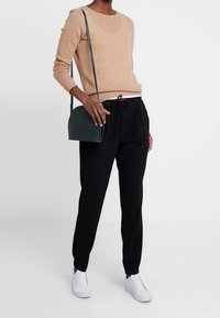Beiger Cashmere-Pullover, schwarze Jogginghose mit Kordelzug und weiße Sneakers. Die Accessoires umfassen eine kleine grüne Schultertasche.