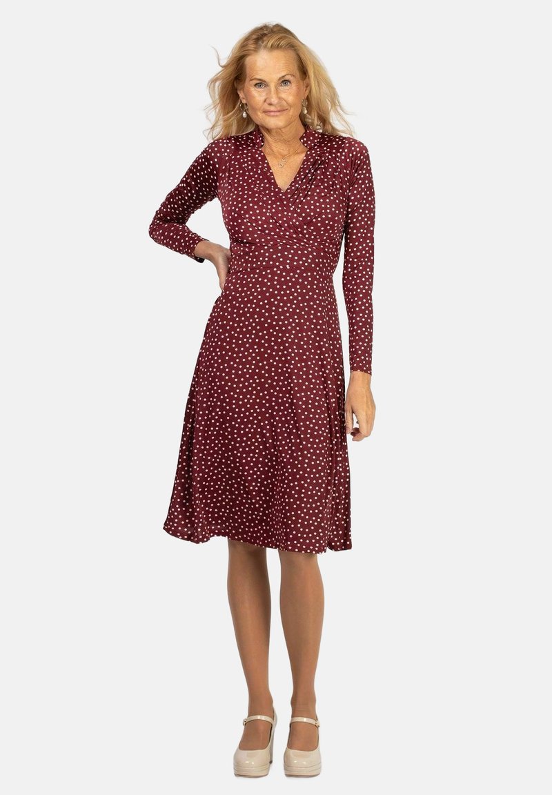 Viininpunainen, pitkähihaiseksi mekko, jossa on valkoisia pilkkuja, varustettuna kietaisukauluksella ja liehuvalla hameella. Mallilla on päällä vaaleat platform-kengät.