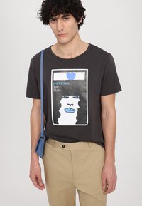Zwarte katoenen t-shirt met een grafische print van een gezicht met blauwe lippen en ogen, gecombineerd met beige broek en een blauwe schoudertas.