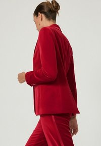 Rød fløjl blazer med struktureret design, enkeltknaplukning og skræddersyet pasform, stylet med matchende røde bukser for et sammenhængende look.