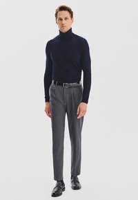 Granatowy sweter z golfem o dopasowanym kroju, zestawiony z szarymi eleganckimi spodniami i czarnymi skórzanymi butami, stojący na białym tle.