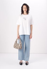 Λευκό βαμβακερό t-shirt με γραφικό "Ζωδίο", συνδυασμένο με ανοιχτό μπλε παντελόνι wide-leg με ασημί σταγονίτσες, κρατώντας μια μπεζ τσάντα από φίδι.