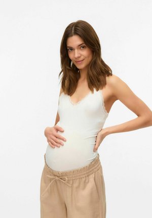 Débardeur de maternité en tissu blanc doux avec des finitions en dentelle, associé à un pantalon beige à taille élastique. Le mannequin serre doucement son ventre rond.