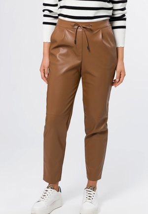 Femme portant un pantalon en similicuir marron avec taille à cordon, des baskets blanches et un pull blanc à rayures horizontales noires.