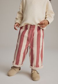 Pantalons rayés en coton doux rouge clair et crème avec une taille élastique et des poignets retroussés, associés à des chaussures beige à enfiler.