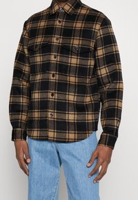 Camicia in flanella a quadri con colori nero e beige, due tasche sul petto e chiusura a bottoni. Il tessuto ha una consistenza morbida e una vestibilità comoda.