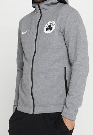 Person trägt eine graue Zip-up-Jacke mit schwarzem Reißverschluss, Celtics-Logo und weißem Nike-Swoosh auf der Brust, Hände in den Taschen der schwarzen Hose.
