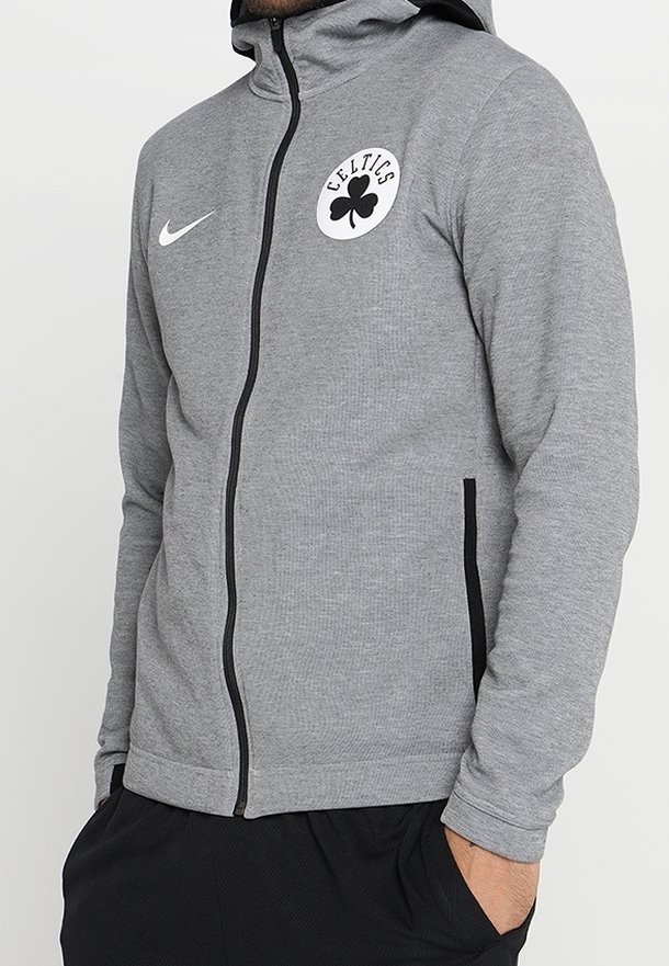 Personne portant une veste grise à fermeture éclair noire, logo des Celtics et swoosh blanc Nike sur la poitrine, mains dans les poches du pantalon noir.