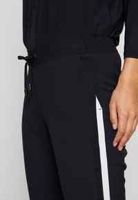 Schwarze Jogginghose mit elastischem Bund und Kordelzug. Besitzt einen weißen Seitenstreifen und eine leichte, glatte Stoffstruktur.