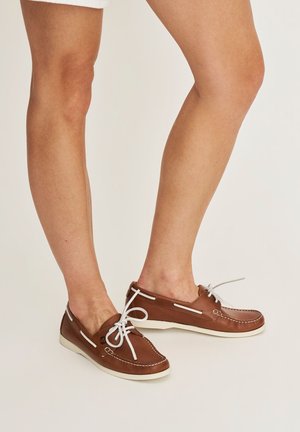 Benen met bruine leren bootsschoenen met witte veters en zolen, gecombineerd met witte shorts tegen een effen achtergrond.