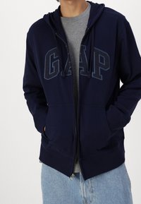Sudadera con cremallera en color marino con el logo "GAP" en un tono más oscuro, cuenta con capucha ajustable, bolsillos delanteros y puños acanalados. Lleva puesta una camiseta gris.