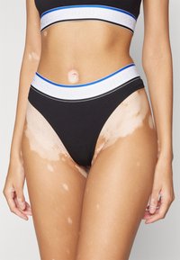 Tanga en coton noir avec une ceinture élastique blanche et bleue portant la marque "HUGO". Texture lisse avec un design ajusté.