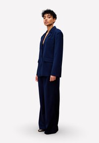 Vrouw staat gekleed in een donkerblauwe oversized blazer en wijde broek met gouden puntige schoenen, kijkt zelfverzekerd vooruit.