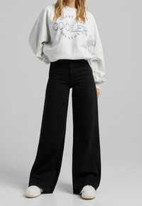 Grå sweatshirt med marinblå text, hög midja svarta vida byxor och vita sneakers på en neutral bakgrund.