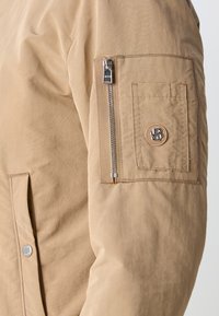 Veste bombers beige avec une texture lisse et mate. Présente une poche zippée sur le bras et une patch ronde avec un logo sur la manche.