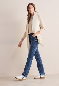 Slim fit blå jeans, krämfärgad stickad tröja och vita sneakers med svarta ränder. Outfiten har en ljus scarf draperad över axlarna.