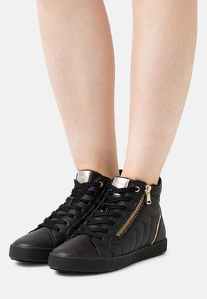 Benen met zwarte hoge sneakers met zwarte veters en gouden ritssluiting op een grijze achtergrond.