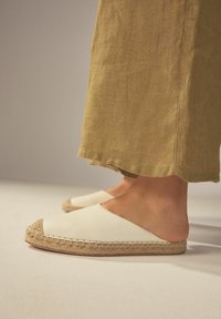 Vita läder slip-on skor med en flätad jute-trim, mot en bakgrund av khaki linnebyxor. Slät textur, öppen design och naturliga material.