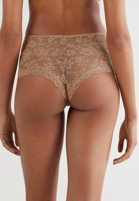 Culotte a vita alta in pizzo di colore nude, con motivi floreali e bordi ondulati, caratterizzate da una texture liscia e un design senza cuciture.