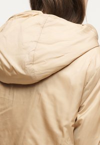 Gros plan d'une personne portant une veste à capuche beige clair, vue de dos avec des cheveux bruns visibles au-dessus du col.