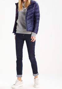 Tmavě modrá prošívaná bunda, šedá mikina s logem, tmavé džíny a světlé tenisky. Součástí jsou kapuce a ohrnuté manžety na džínách.