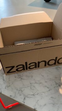 Brown cardboard box partially open with the Zalando logo in black. Inside, a white item is visible, resting on a white marble surface.