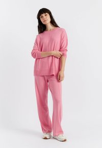 Pull en maille rose clair à la coupe décontractée et fentes sur les côtés, associé à un pantalon rose assorti ; mannequin portant des baskets blanches.