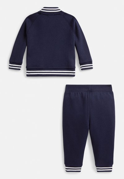 Hanorac albastru închis cu dungi albe pe manșete și tiv, asortat cu pantaloni conici cu talie elastică și accente dungi.