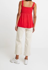 Röd smocktop med breda axelband, matchad med krämfärgade vida byxor. Modellen bär vita sneakers, vilket visar en avslappnad outfit.