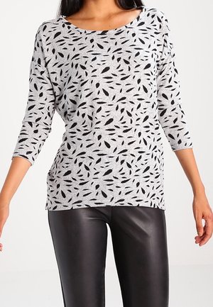 Femme portant un haut gris à manches longues avec des motifs de feuilles noires et un pantalon en cuir noir sur un fond uni.