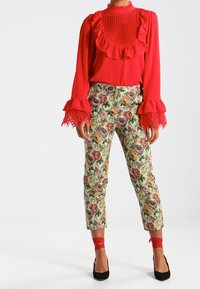 Blusa rossa con scollatura a rouches e polsini rimborsati, abbinata a pantaloni a fantasia floreale verdi, con dettagli rossi e tacchi neri.