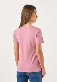 Maglietta rosa a maniche corte in cotone con scollo rotondo e taglio aderente, vista da dietro, abbinata a jeans blu. Tessuto liscio.