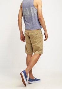 Débardeur bleu clair avec un dos à motifs, shorts beiges et chaussures en toile bleues à enfiler avec semelles blanches. Tenue décontractée vue de dos.