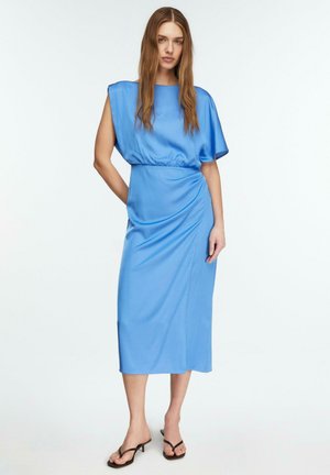 Kobieta stojąca w błyszczącej niebieskiej sukience midi bez rękawów z asymetrycznym wzorem na ramieniu, w czarnych sandałach na obcasie, na białym tle.