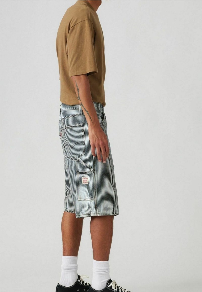 Personne portant une chemise marron à manches courtes et un short rayé bleu et blanc jusqu'aux genoux avec des poches, des chaussettes blanches et des baskets noires.