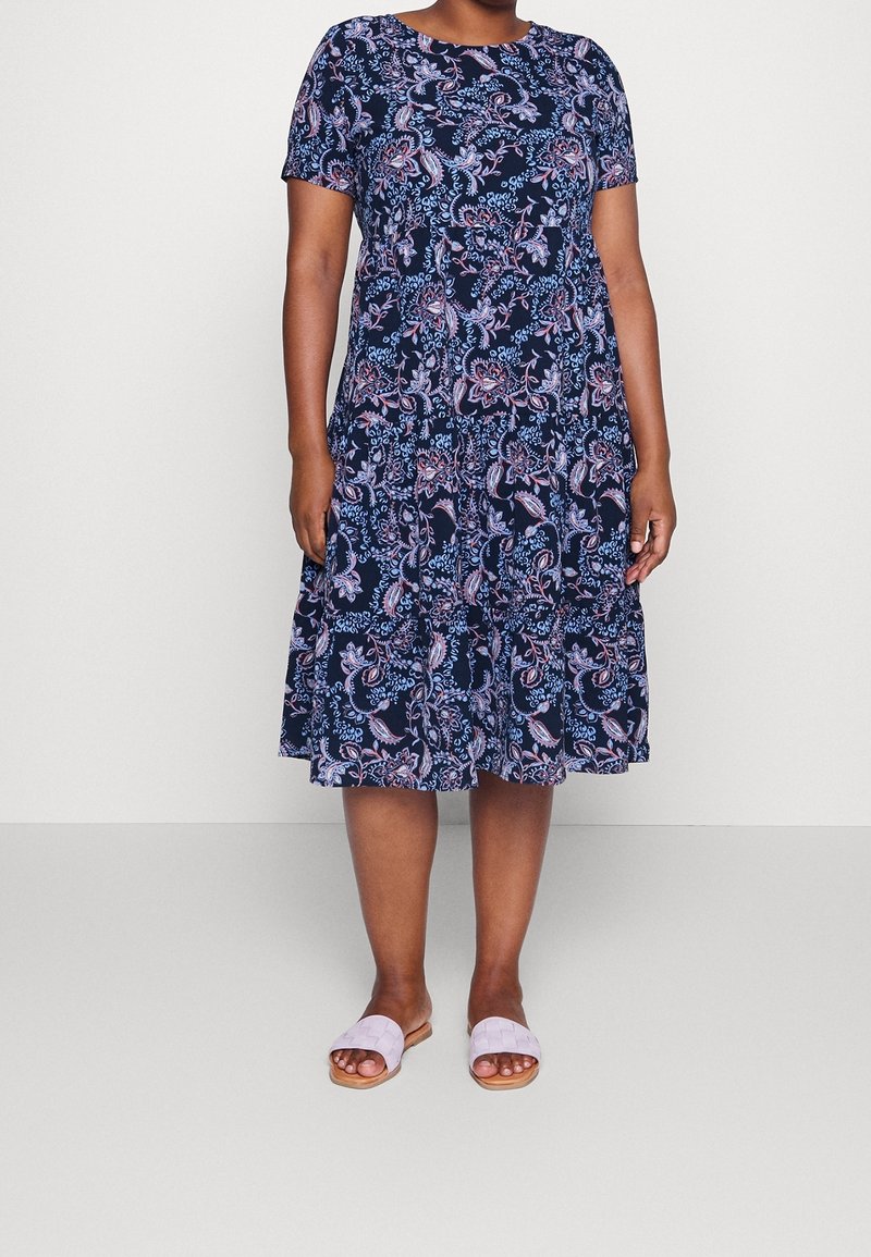 Femme portant une robe midi bleu marine à fleurs, manches courtes, jupe à étages et sandales plates violettes claires sans bride.