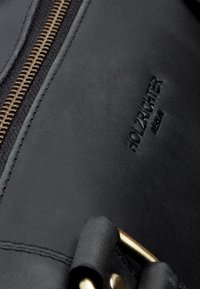 Schwarzer Leder-Rucksack mit einem goldfarbenen Reißverschluss und geprägt Logo. Besitzt eine glatte Textur und dezente Nähdetails.